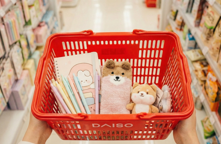 A red Daiso shopping basket filled with cute Korean stationery, pastel pens, a character notebook, and fluffy animal socks in a store aisle.